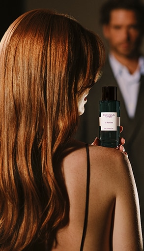 Hosszú, fényes vörös haj hátulnézetben, kézben tartott Hair Rituel by Sisley Le Parfum luxus haj- és fejbőrápoló illat üvegével.