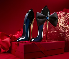 Carolina Herrera Good Girl parfüm stiletto formájú üvegek fekete masnival, piros ajándékdobozok és elegáns háttér.
